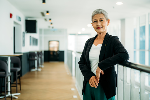 Confident stylish Asian mature middle aged woman standing at modern workplace. Stylish older senior businesswoman, 60s gray-white haired lady executive leader manager looking at camera in office, portrait.
