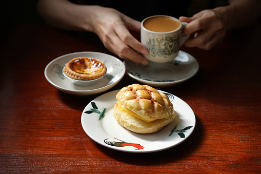 Cropped shot shows a young Asian woman enjoying freshly baked Hong Kong style polo bun and egg tart with a cup of milk coffee at a local tea restaurant.