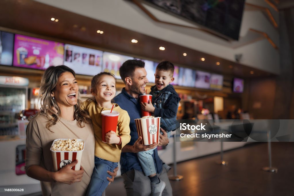 Young happy family with popcorn and drinks in movie theatre. Happy family enjoying while walking with popcorn and drinks in cinema. Movie Theater Stock Photo