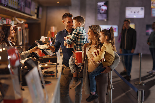 Happy parents and their kids buying movie tickets at concession stand in cinema.