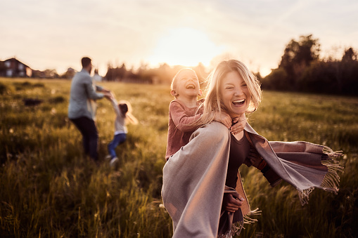 Carefree woman having fun while piggybacking her small son in the park at sunset. There are people in the background. Copy space.