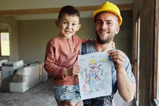 Happy manual worker and his small son holding a drawing during home renovation process and looking at camera.