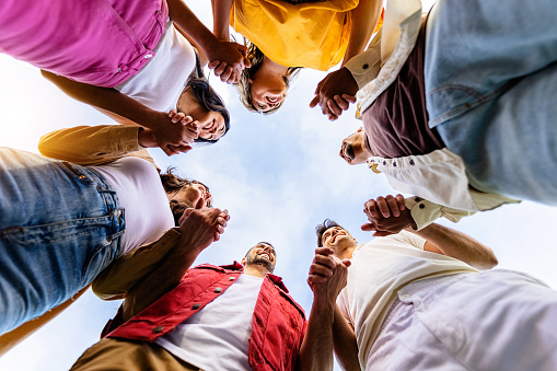 Low angle view of united group of young people in circle holding hands. Community, support and teamwork concept with millennial people showing unity outdoors.