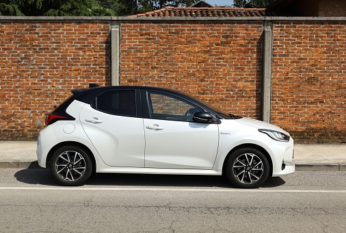 Udine, Italy. May 6, 2023. White and black Toyota Yaris hybrid at the road side. Side view, brick wall on background