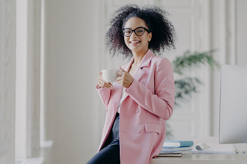 Photo of relaxed businesswoman holds cup of hot drink, has coffee break, stands near her workplace in spacious white cabinet wears spectacles long pink jacket works in office. Time for rest after work