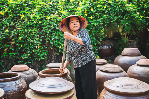 portrait of vietnamese senior woman with straw hat stirring soy sauce in huge clay pod standing in a row in courtyard