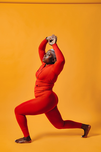 Portrait of mid adult black plus size woman wearing red jumpsuit with arms raised, lunging, in yoga pose against yellow studio background