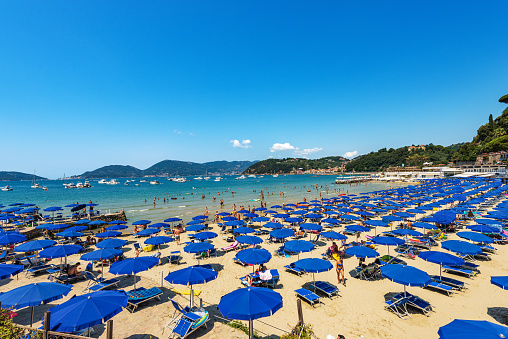 Lerici, Italy -July 16, 2022: Beach of Lerici Town with many umbrellas, tourist resort on the coast of the Gulf of La Spezia or Gulf of Poets, Liguria, Italy, Europe. On background the small village of San Terenzo and Porto Venere. A large number of tourists relax or sunbathe on the beach and swim in the bay on a sunny summer day.
