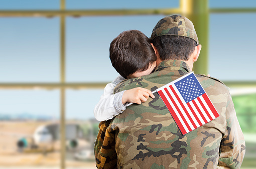 Shot of an adorable child hugging his father affectionately with the united states flag in his hand with a blue sky and copy space.