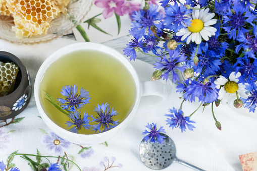 Cornflower herbal tea in white cup on white crochet napkin intdoors, healthy cornflower drink with honey, fresh cornflowers flowers in vase, healthcare and healthy eating concept