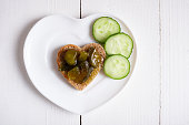 Cucumber jam is spread on bread in the shape of a heart next to a fresh sliced cucumber. The sandwich lies on a bowl in the shape of a heart