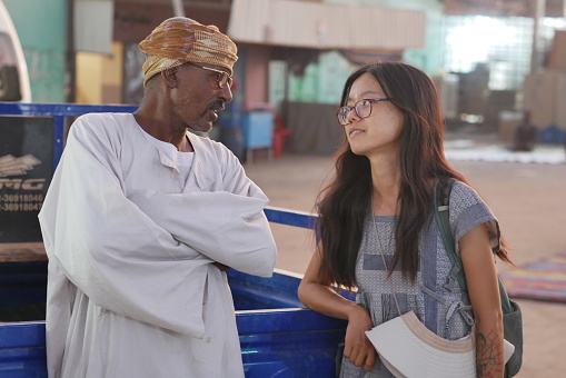 Wadi Halfa, Sudan – 10.16.2022: Pretty female Chinese tourist without hijab chats with local Sudanese man in traditional dress