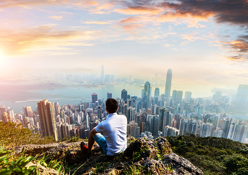 Young man sitting on top of mountain and looking at hong kong skyline