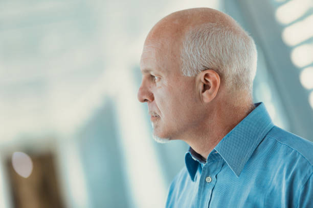 After so many years, you never know if it will end the same way. Profile view of old man in office attire looking between illuminated slits in a modern corridor of a hospital or business. He looks out and seems to be checking. never the same stock pictures, royalty-free photos & images