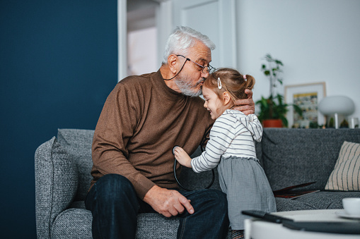 Portrait of a senior man playing with his granddaughter. He is sitting on the sofa while she is using stethoscope to act as a doctor or a nurse. They are smiling and enjoying their time with each other.