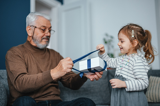 Portrait of a senior man playing with his granddaughter. He is sitting on the sofa wearing glasses and looking down at the gift box she brought for him to open. They are both smiling.