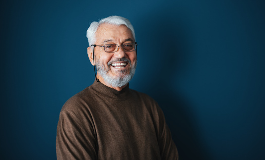 Close up shot of a handsome senior man wearing glasses posing by the blue wall. He is wearing a brown sweater and there is dark blue wall behind him. He is smiling and looking away.