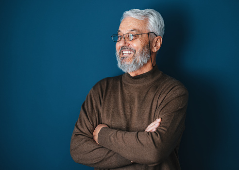 Close up shot of a handsome senior man wearing glasses posing by the blue wall. He is wearing a brown sweater and there is dark blue wall behind him. He is smiling and looking away.