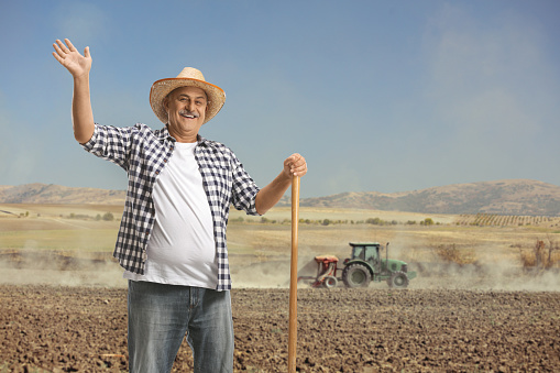 A farmer standing outdoors, leaning against the wooden wall of a red barn, wearing overalls and a cowboy hat. He is standing with a serious expression on his face, looking toward the camera. He is a senior man in his late 60s.