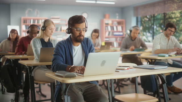 Diverse Mature Students Learning in Classroom, Sitting Behind Desks with Headphones, Using Laptops and Writing in Notebooks. Teacher Giving an Adult Education Course Remotely Online