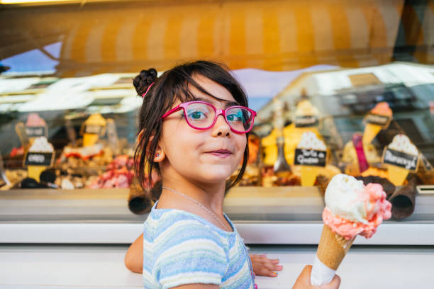 Girl with glasses eating an ice cream in front of an ice cream shop looking at camera stock photo