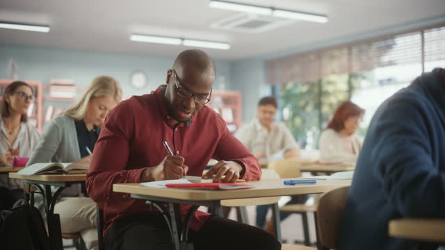 Young Black African Man Raising Hand and Asking Teacher a Question During a Test in Adult Education School. Diverse Mature Students Learning in Classroom, Writing Exam Answers in Notebooks
