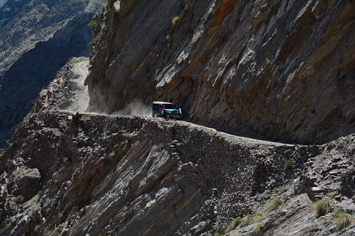 A sturdy jeep navigates a treacherous road, carved between towering mountains. The rugged terrain presents a daunting challenge as the vehicle maneuvers through sharp bends and steep inclines.