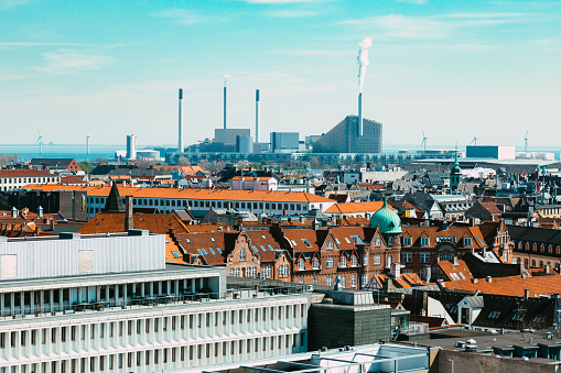 High angle view of the urban skyline of Copenhagen, Denmark, with the power plant of Copenhill emitting its fumes into the sky. Located in Amager in Copenhagen, CopenHill is a waste-to-energy plant topped with a ski slope, a hiking trail, and a climbing wall. The power plant plays a part in Copenhagen's goal of becoming the world's first carbon-neutral city by 2025, combining an architectural landmark with an urban recreation center.
