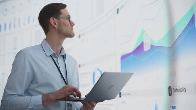 Close Up Of Successful Caucasian Man Typing On Laptop Computer And Standing Next To Big Digital Wall With Data In The Office. Male Business Analyst Creating A Growth Strategy For Innovative Startup.