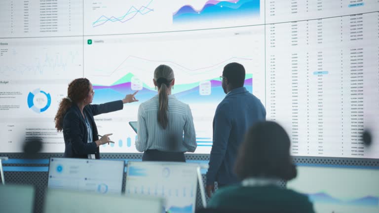 Diverse Team Of Data Scientists Having A Meeting And Discussing Reports In a Modern Monitoring Office With Analytics Feed On a Big Digital Screen. Deep Learning Company With Employees Behind Computers