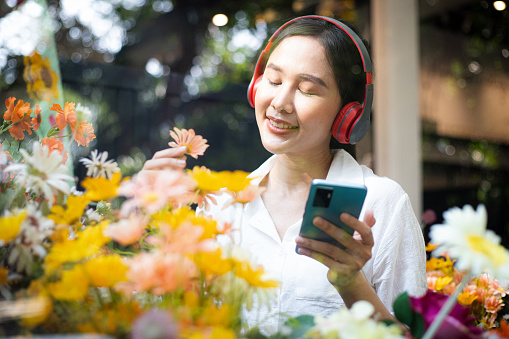 Portrait of smiling woman smelling sunflowers at flower shop with music on smartphone. Florist in the modern house in sunny day.