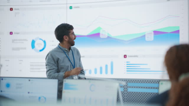 Male Indian Computer Scientist With Tablet Giving Presentation to Diverse Team Of Data Analysts In Front Of Big Digital Screen In Monitoring Room. Colleagues Listening To Training On Automation.