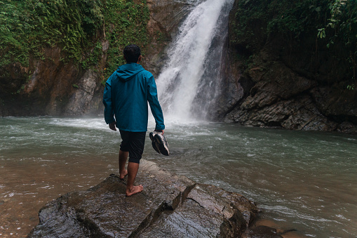 an asian man on barefoot enjoying a scenic view of a waterfall while holding his shoes in a tropical rainforest