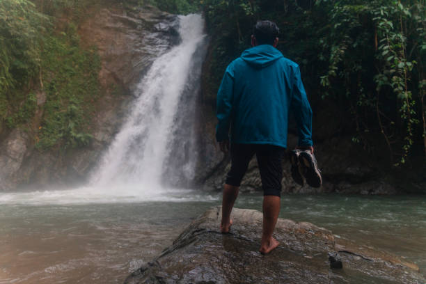 an asian wan enjoying a scenic view of a waterfall while holding his shoes in a tropical rainforest stock photo