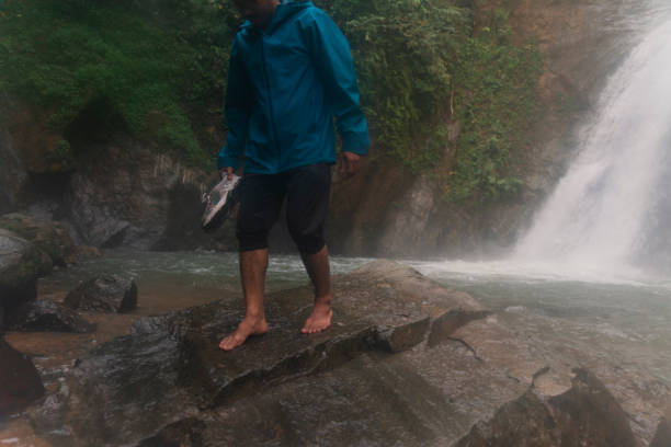 an asian man on barefoot walking down on stone with waterfall scene in the background in a tropical rainforest stock photo