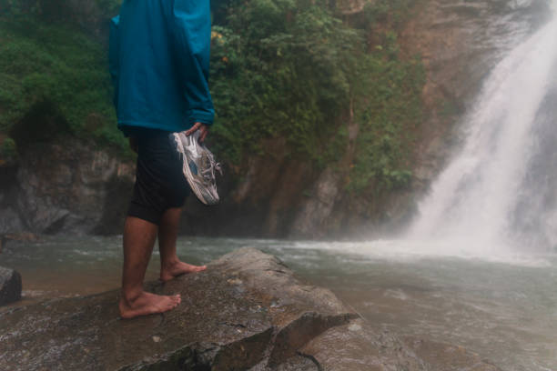 an asian wan enjoying a scenic view of a waterfall while holding his shoes in a tropical rainforest stock photo