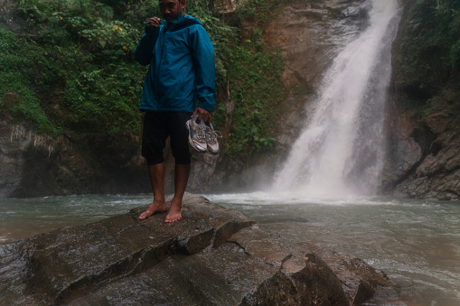 an asian man barefoot walking down on stone with waterfall scene in the background in a tropical rainforest