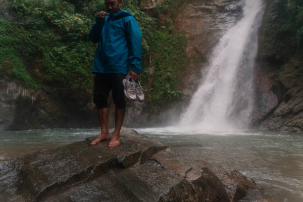an asian man barefoot walking down on stone with waterfall scene in the background in a tropical rainforest stock photo