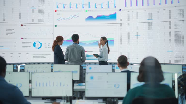 Wide Back Shot of Diverse Team of Data Scientists Discussing Graphs And Reports On Big Digital Screen In Monitoring Office. Multiethnic Employees Working On Desktop Computers For Deep Learning Company