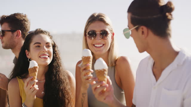 Summer, ice cream and friends outdoor at beach talking, happy and eating cones for fun. Young men and women group lick vanilla gelato dessert while laughing for travel adventure, holiday or vacation