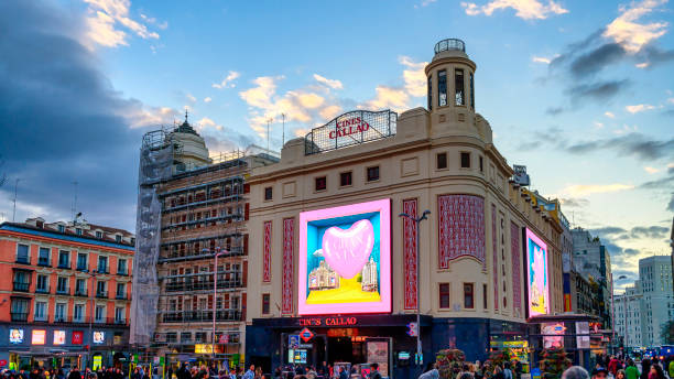 Madrid, Spain - February 8, 2023: A modern high-rise building with small towers on its rooftop surrounded by other buildings. A crowd of people is visible in the foreground at dusk with a cloudy dark blue sky above.