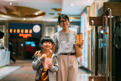 Modern asian woman and her son holing cup of fizzy drink and bucket of popcorn walking to cinema hall to watch movie.