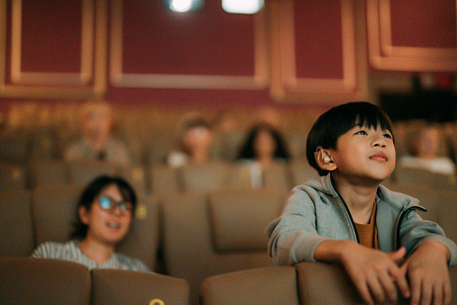 A mother sits back while her son leans forward with interest to watch a movie at the cinema.
