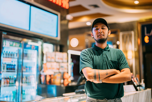 Portrait of movie theater asian man staff looking at camera.An Asian male theater employee stood arm crossed in front of a beverage machine, smiling and looking at the camera.