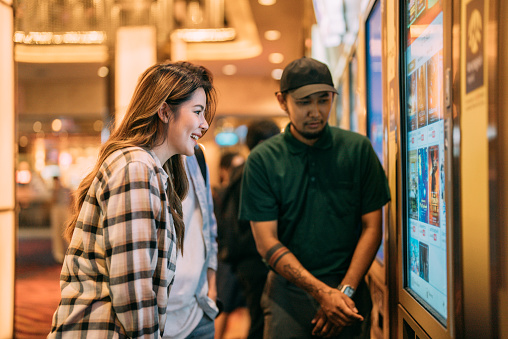 Asian young lover choosing movie and buying ticket from vending machine at movie theater at mall. Young woman making gestures by touching screen
