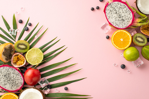 Tropical getaway. A stunning top view flat lay of exotic fruits like dragon fruit, kiwi, orange, lime, tomarillo, carambola and coconut, palm leaves on a pastel pink background, with a copyspace