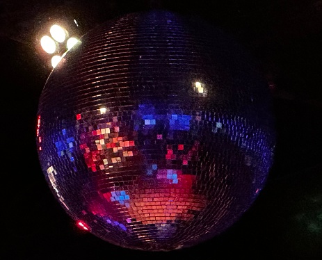 A hanging disco ball reflects dark shades of blue, purple, red, and orange.Four bright lights are visible, offsetting the black background.