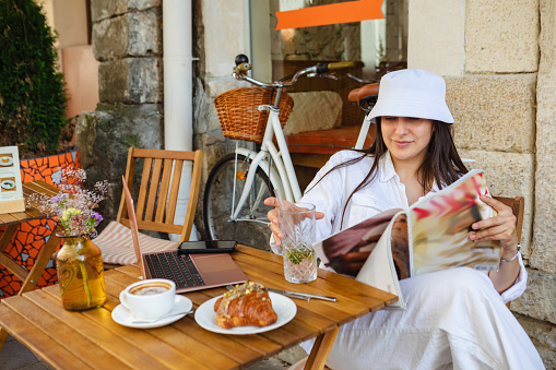 girl with glass of lemonade and magazine sits at a table in a cafe with a laptop on it