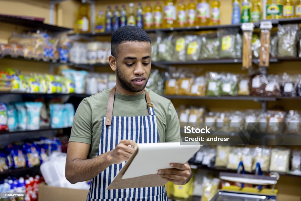 Grocer working at a local food shop doing an inventory African American grocer working at a local food shop doing an inventory and holding a clipboard Convenience Store Stock Photo