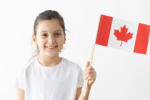 Little girl is holding Canadian flag in hand and is looking at camera in front of white background.